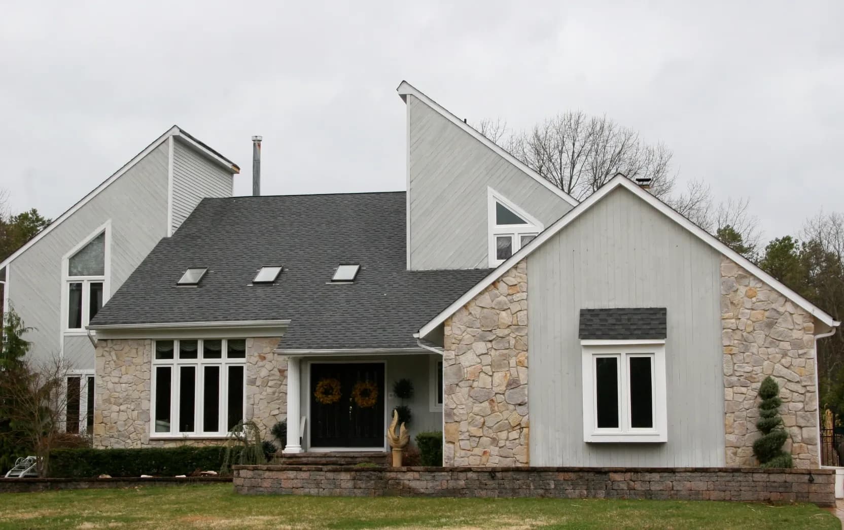 Modern house with a mix of stone and wood siding, featuring sharp angles and large windows. Overcast sky, neatly trimmed shrubs, and grassy lawn.