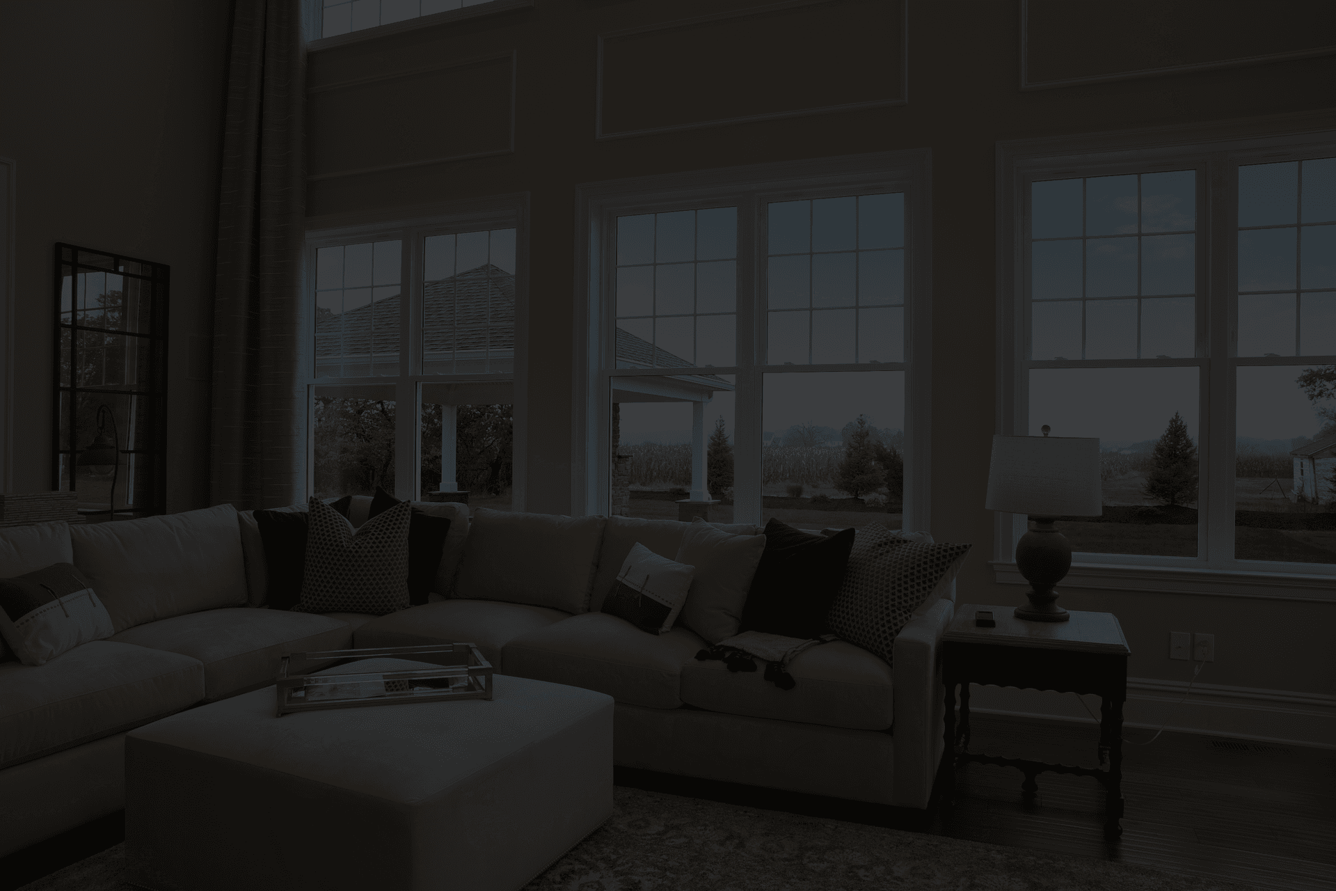Spacious living room with a large beige sectional sofa, assorted green and beige pillows, and a cushioned ottoman. Bright windows offer a view of a garden and sky. A table with a lamp adds warmth.