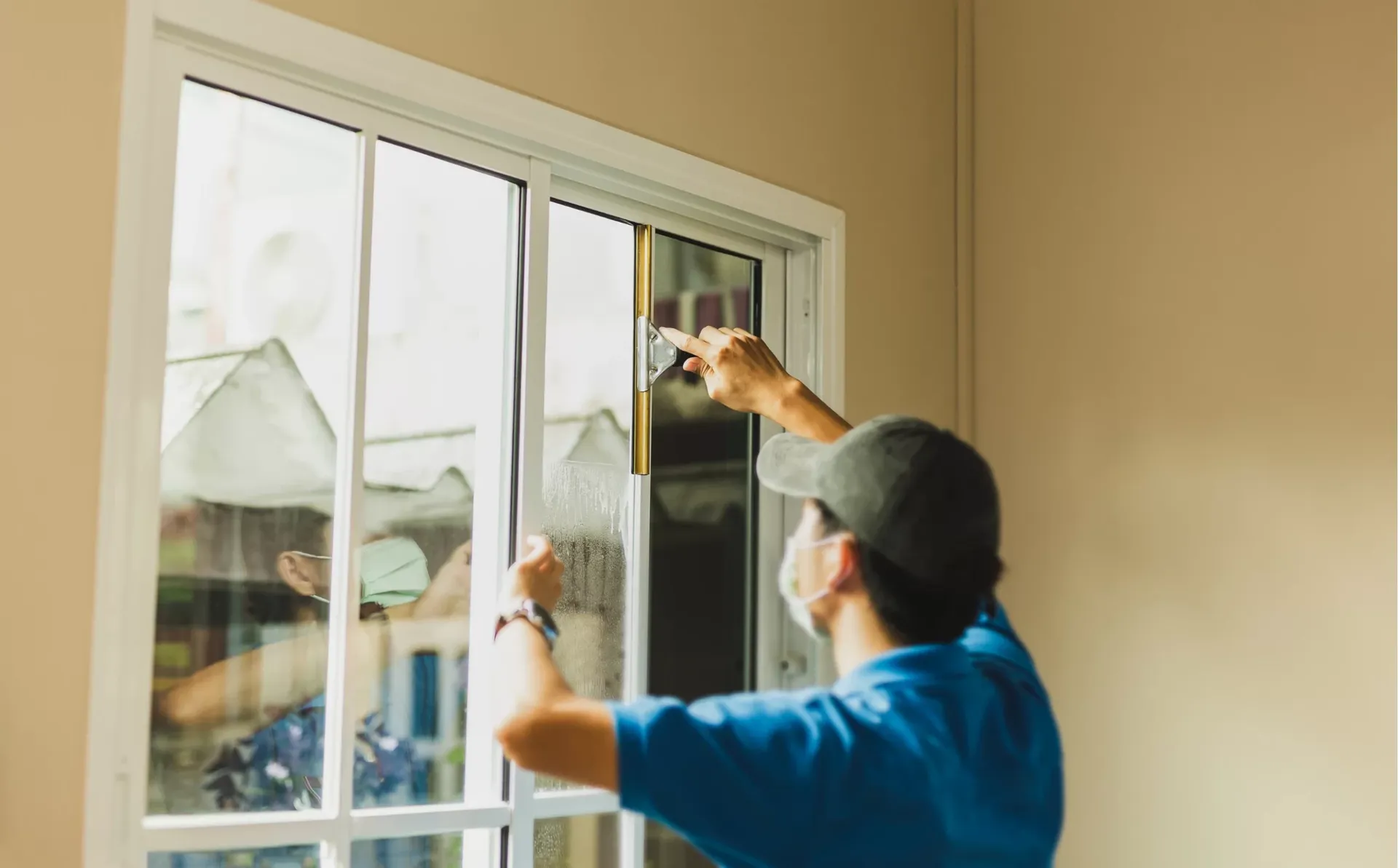 A person in a blue shirt and cap is cleaning a glass sliding door using a scraper. They are wearing a mask, focusing intently, with bright light streaming in.
