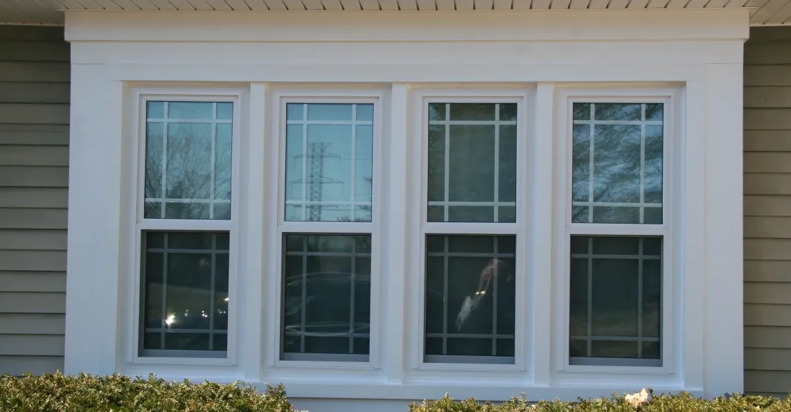 A large white-trimmed triple-pane window on a beige house exterior. The window reflects cloudy skies and trees, with green bushes lining the base.